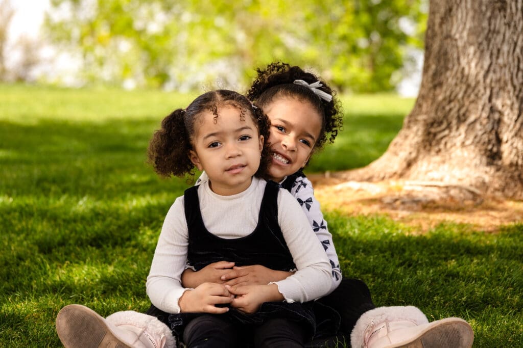 Two young sisters sitting together on the grass beneath a tree at Anthem Community Park